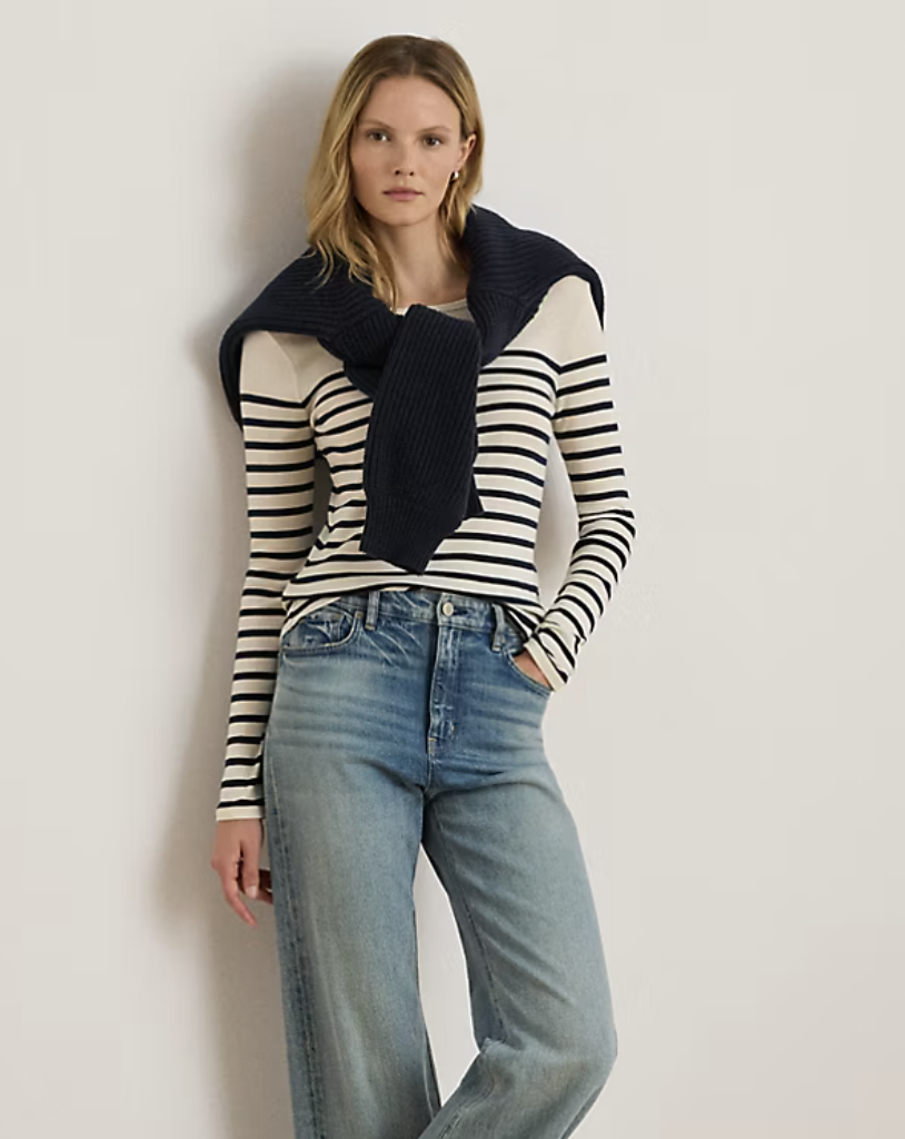 Woman wearing striped long-sleeve top with navy sweater draped over shoulders and jeans demonstrating versatile travel outfit styling